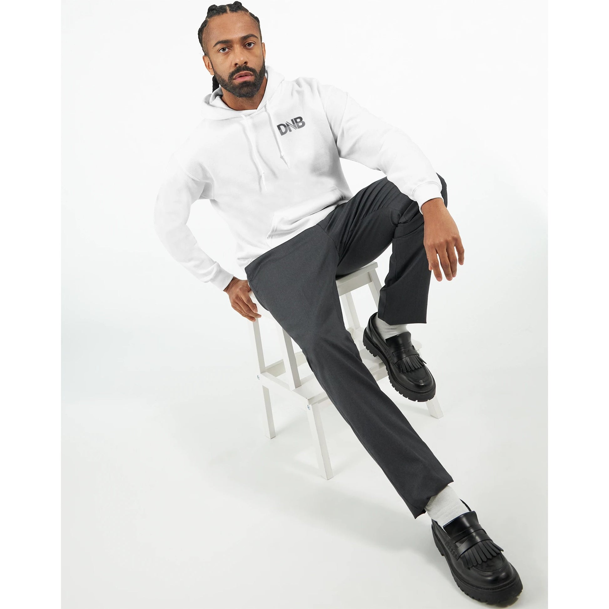 Man sitting on a stool wearing a white hoodie with text, gray pants, and black shoes on a white background